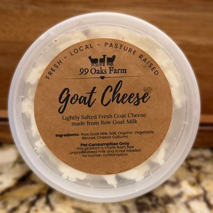A round plastic tub of 99 Oaks Farm Goat Cheese with a brown paper label, set against a blurred wooden and stone background.