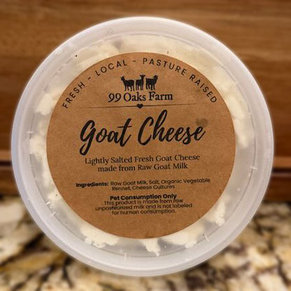 A round plastic tub of 99 Oaks Farm Goat Cheese with a brown paper label, set against a blurred wooden and stone background.